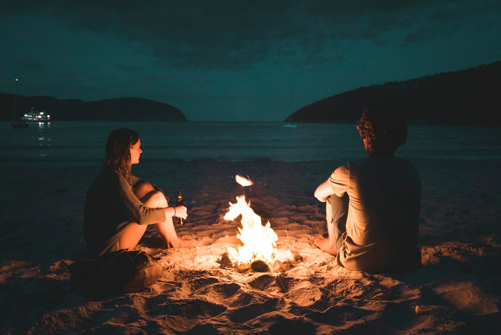 Dos personas conversan en calma junto a una fogata encendida en la playa de noche
